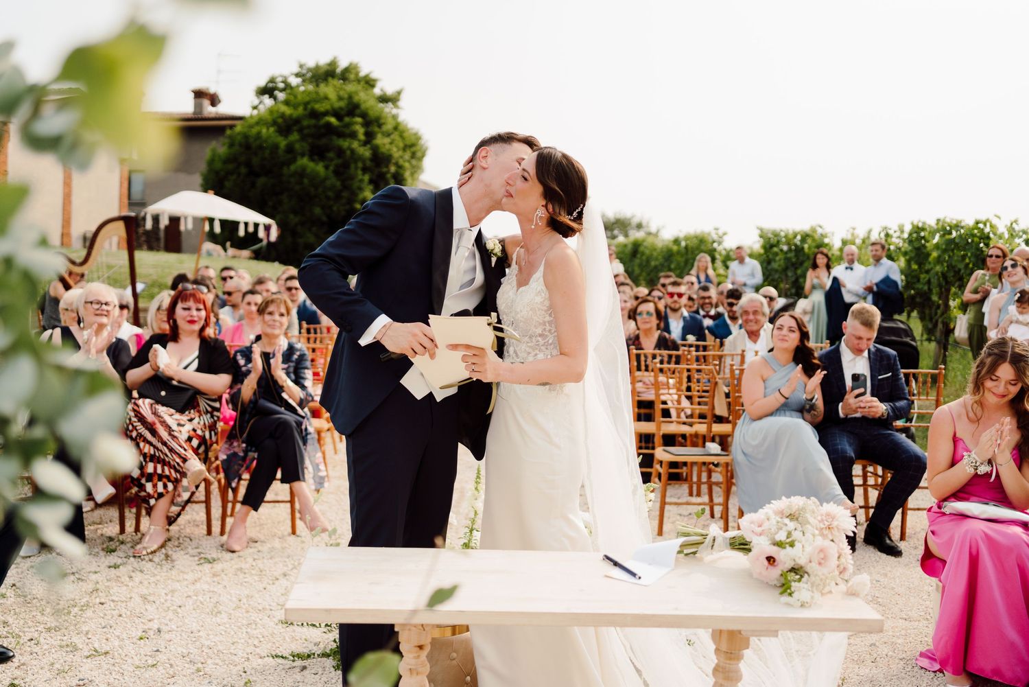 Bride and groom share first kiss at outdoor wedding ceremony while guests applaud.