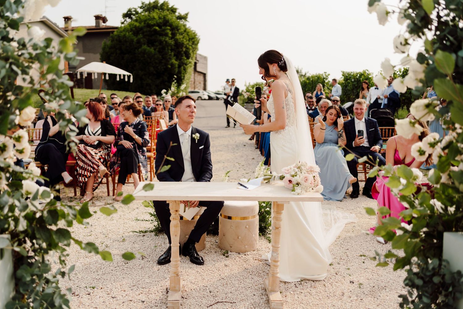 Bride and groom at signing table during outdoor wedding ceremony framed by greenery.
