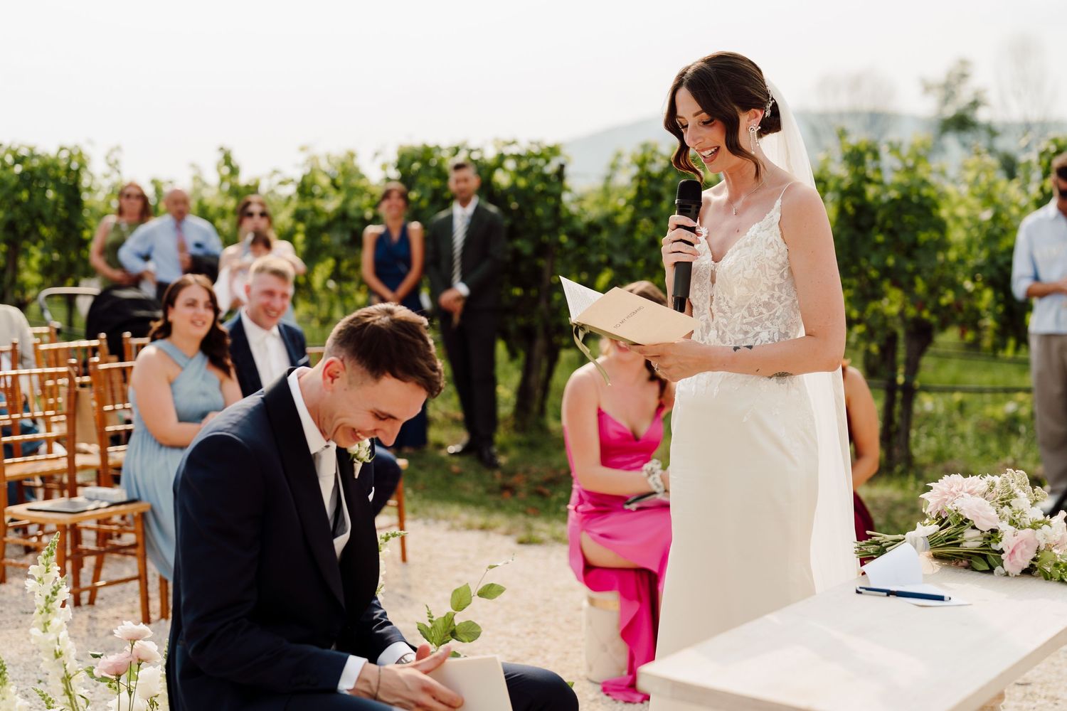 Bride reads vows at microphone while groom signs documents at outdoor wedding ceremony.