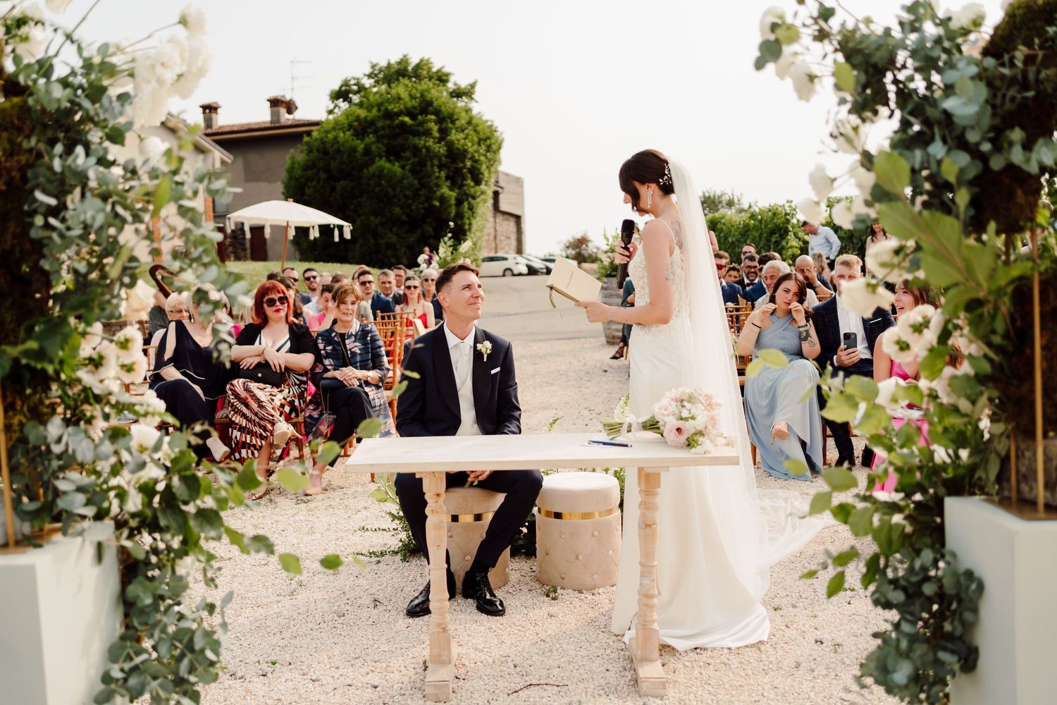 Bride reads vows to seated groom at elegant outdoor vineyard wedding ceremony.