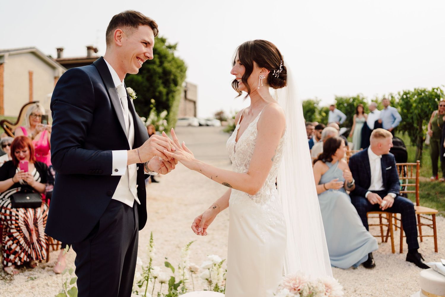 Groom places wedding ring on brides finger during outdoor ceremony with guests watching.