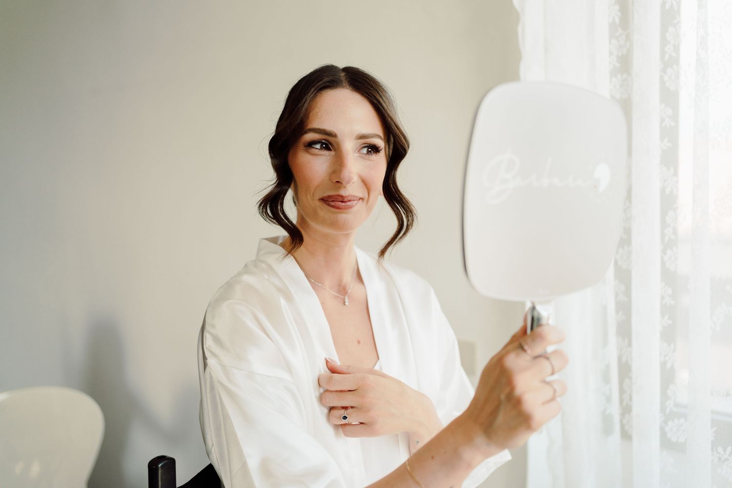 Smiling brunette bride in white silk robe holding a decorative hand mirror during wedding morning preparations.