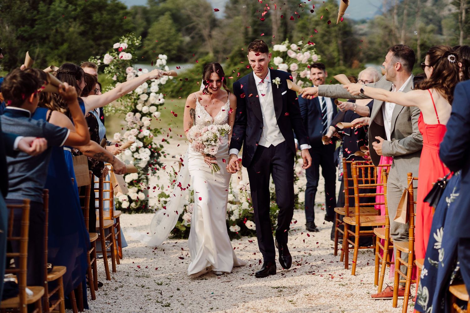 Newlyweds walking down aisle as guests toss confetti at outdoor wedding ceremony.