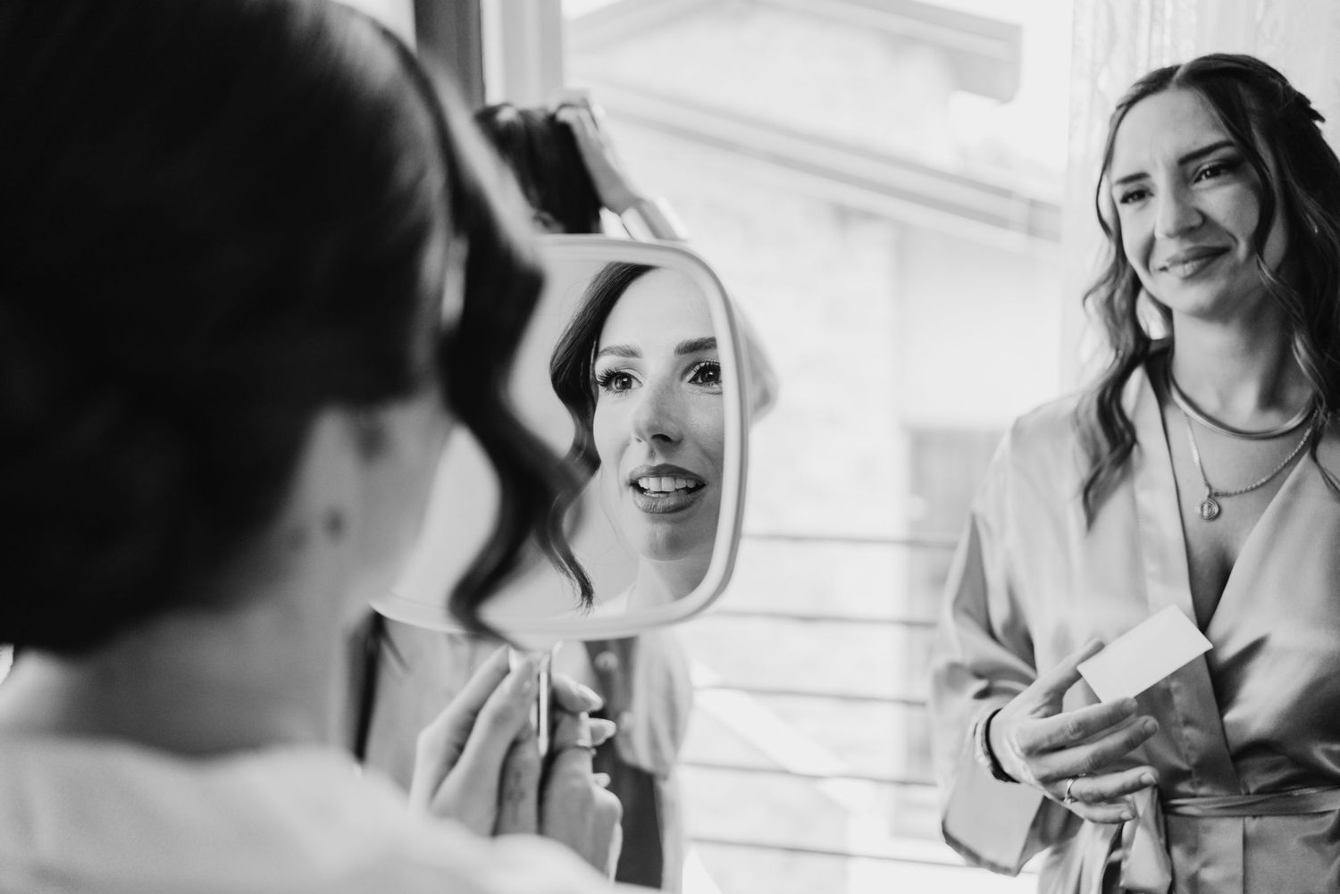 Black and white photo of bride checking makeup in mirror while bridesmaid smiles beside her during bridal prep.