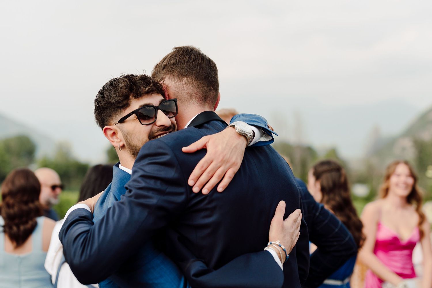 Two men in suits sharing a warm congratulatory hug at outdoor wedding.