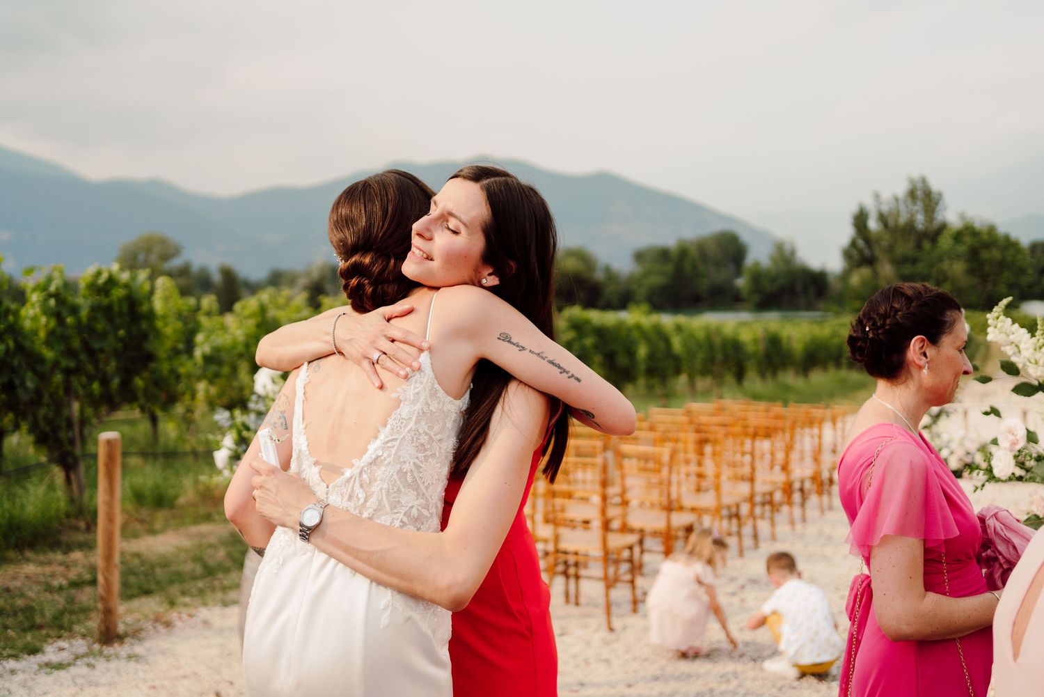 Two women sharing a warm embrace at an outdoor vineyard wedding ceremony with mountain views.