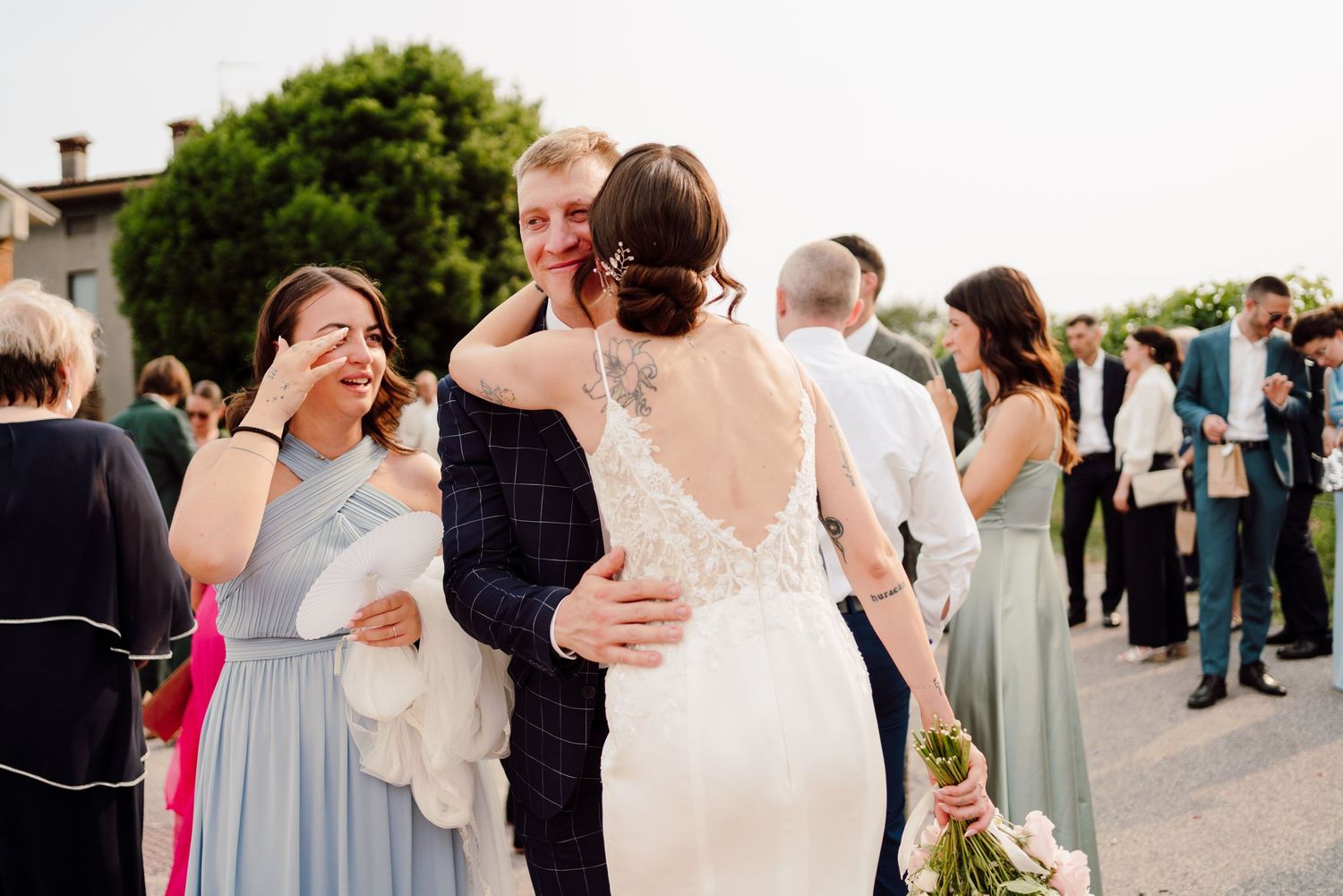 Emotional wedding guest wiping tears while hugging bride and groom outdoors.