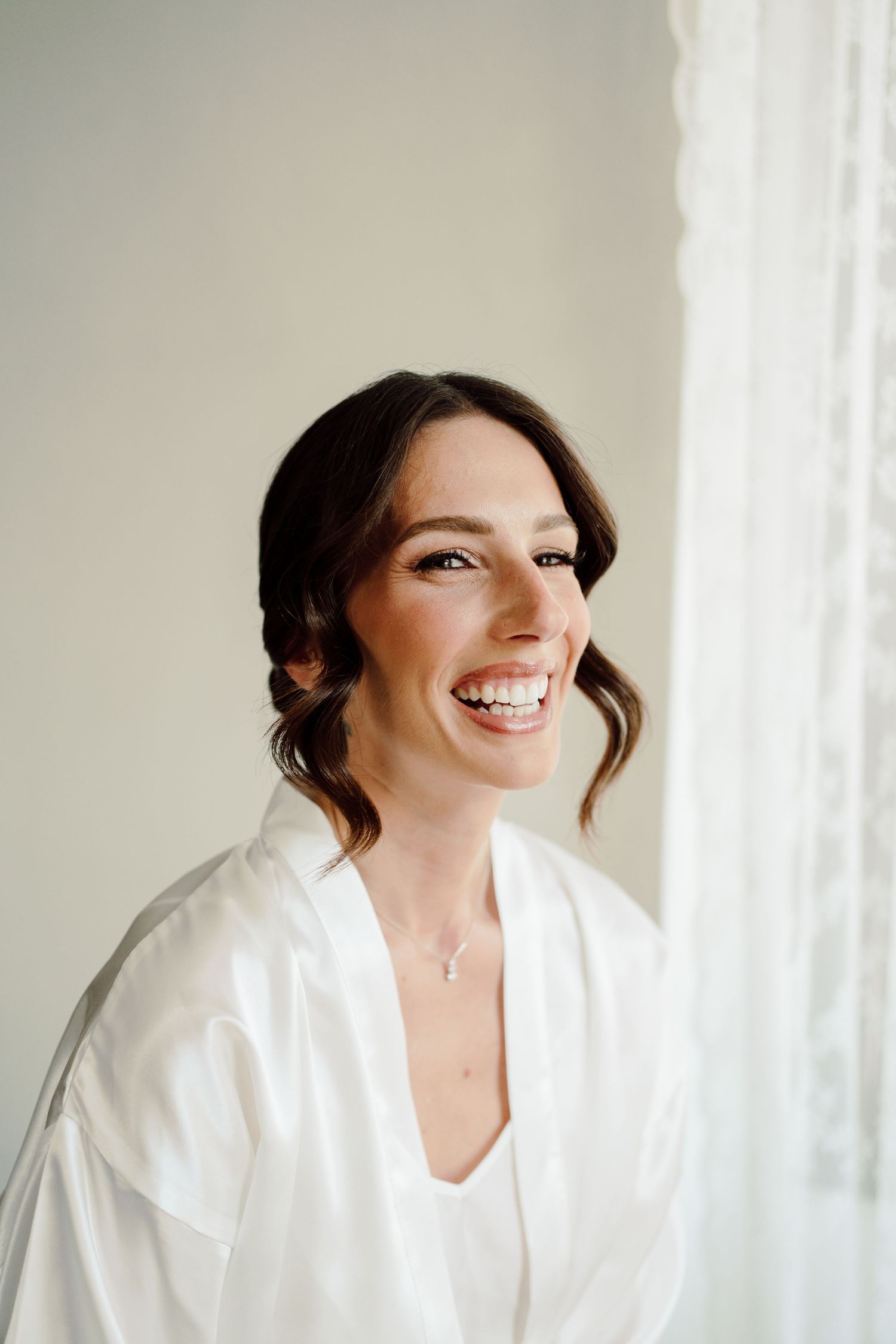 Radiant brunette bride laughing joyfully in white silk robe beside a bright window during wedding morning prep.