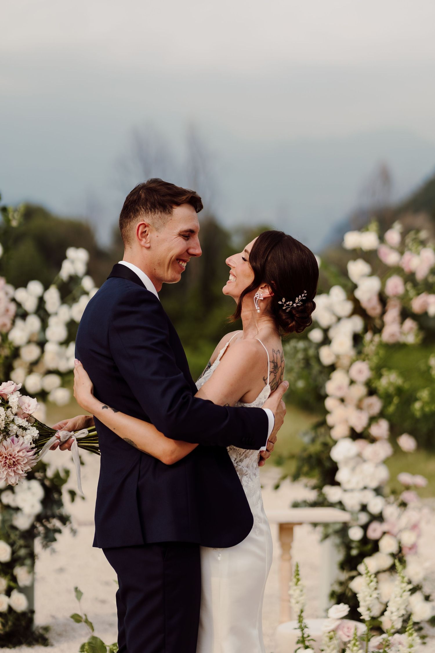 Bride and groom laughing together embracing before floral arch at wedding ceremony.