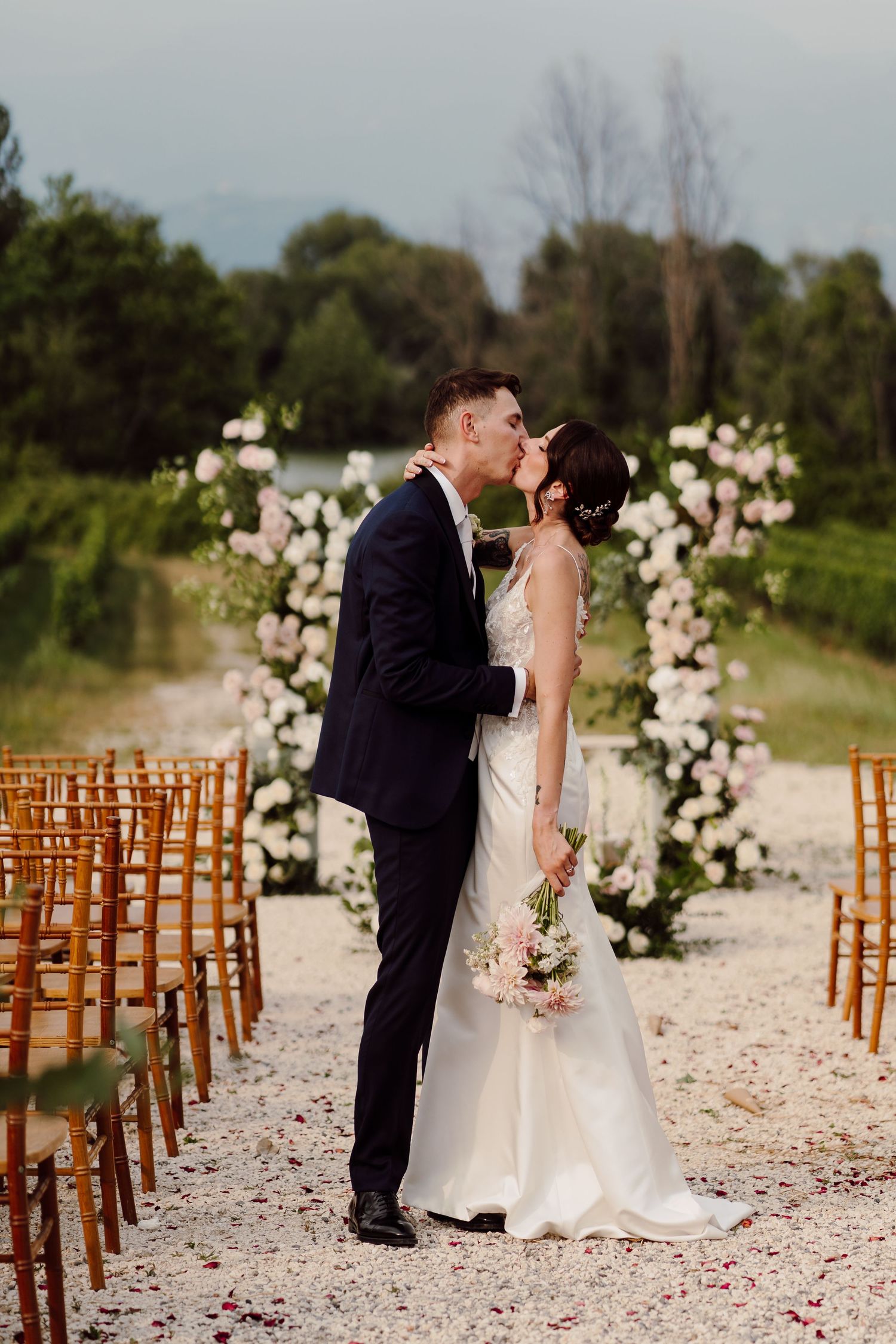Bride and groom sharing their first kiss at an outdoor wedding ceremony lined with wooden chairs.