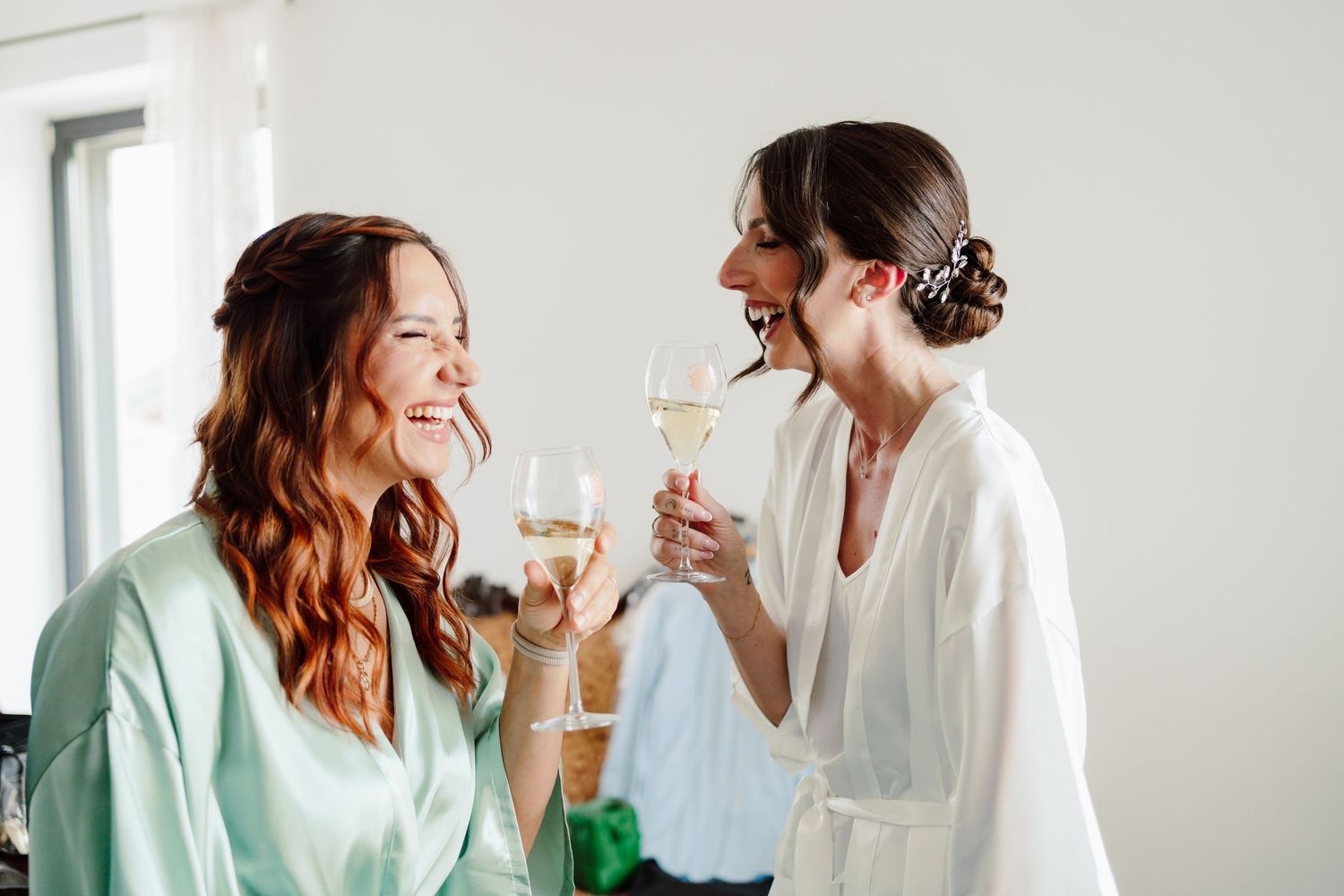 Two joyful bridesmaids in sage and white satin robes toasting with champagne glasses during wedding morning prep.