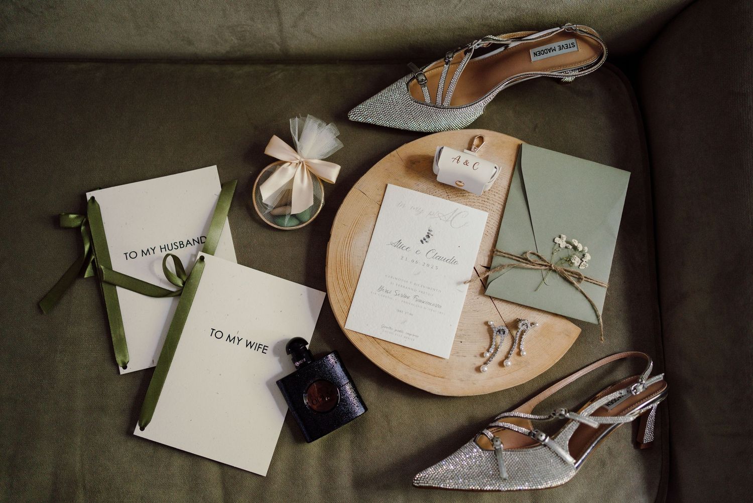 Elegant wedding flat lay with glitter heels, vow books, invitation, perfume, and sage green accents on a velvet surface.