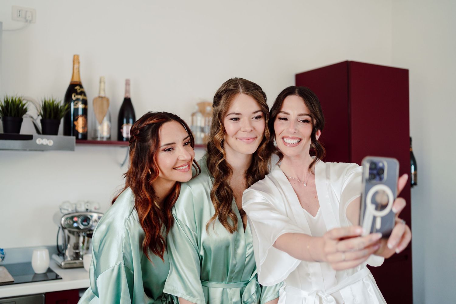 Bride and two bridesmaids in satin robes taking a fun selfie together during wedding morning preparations at home.