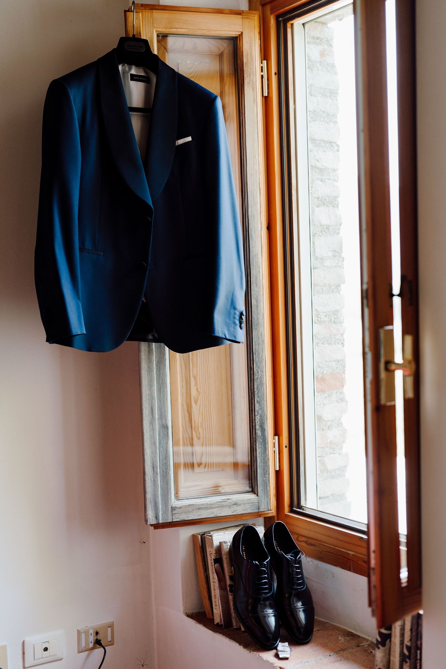 Navy blue wedding suit jacket hanging on a wooden window frame with black Oxford dress shoes below.