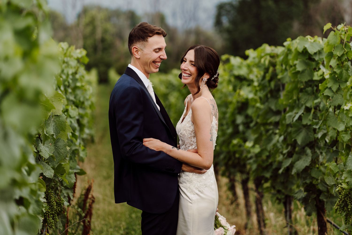 Smiling bride and groom embracing romantically among lush green vineyard rows on their wedding day.