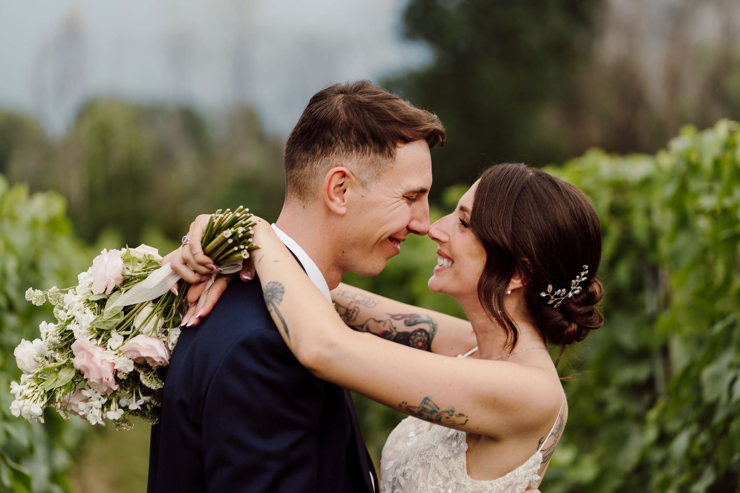 Bride and groom touch noses tenderly in a vineyard, bride holding a lush bouquet.
