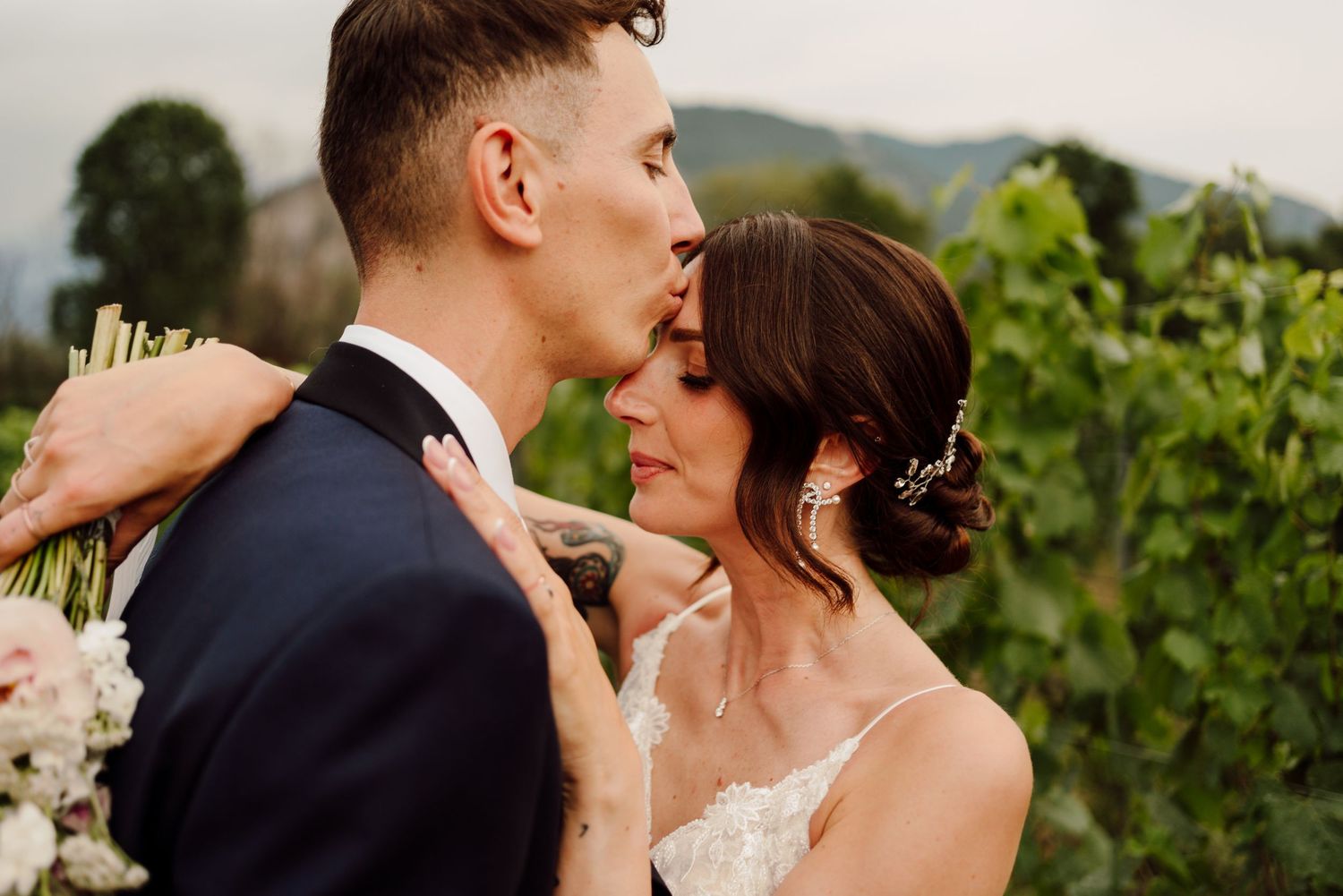 Groom kisses bride on forehead in a vineyard setting on their wedding day.