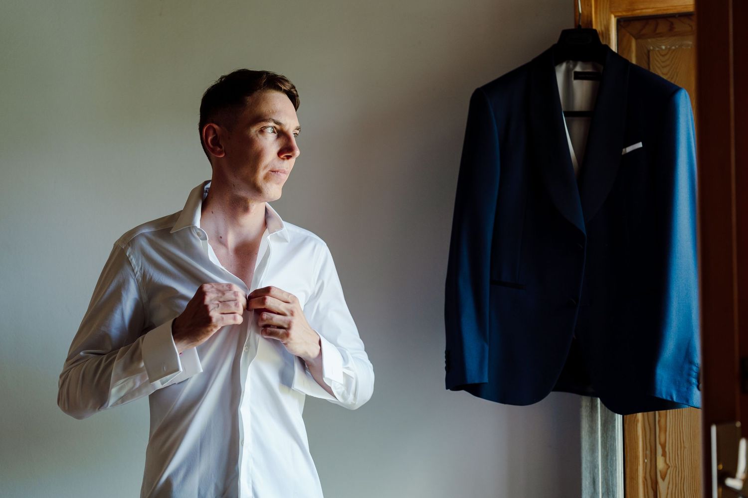 Groom buttoning his white dress shirt before wedding with navy suit jacket hanging in background.