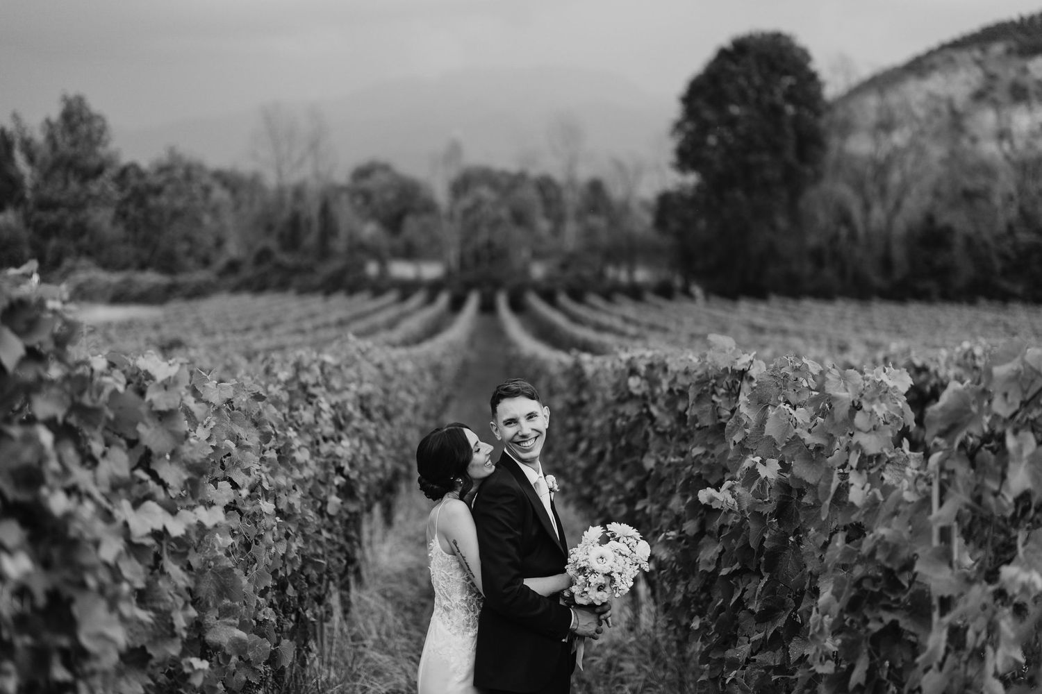 Black and white photo of bride and groom embracing in a scenic vineyard landscape.
