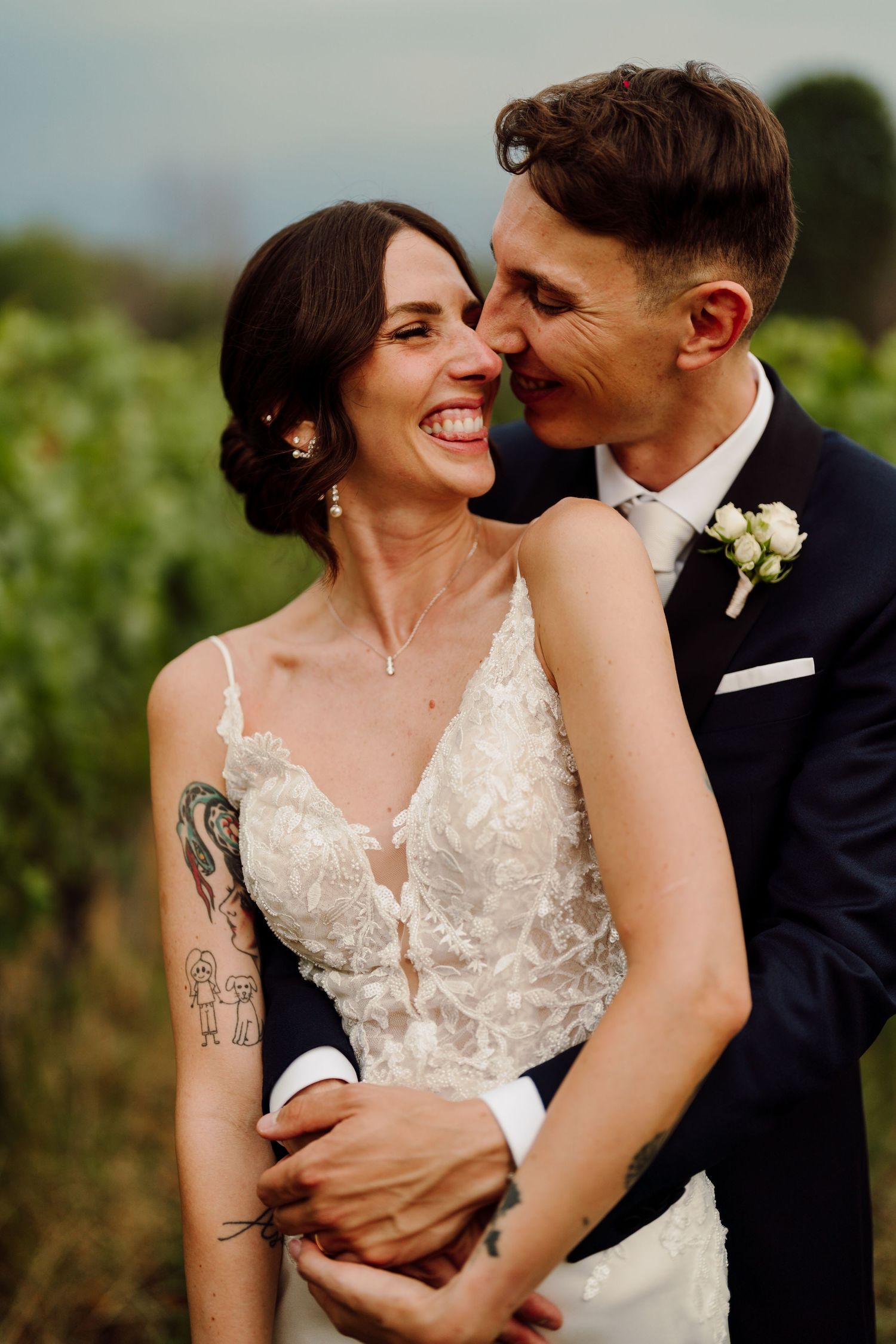 Groom tenderly nuzzling bride with tattoos as she laughs joyfully in a vineyard setting.