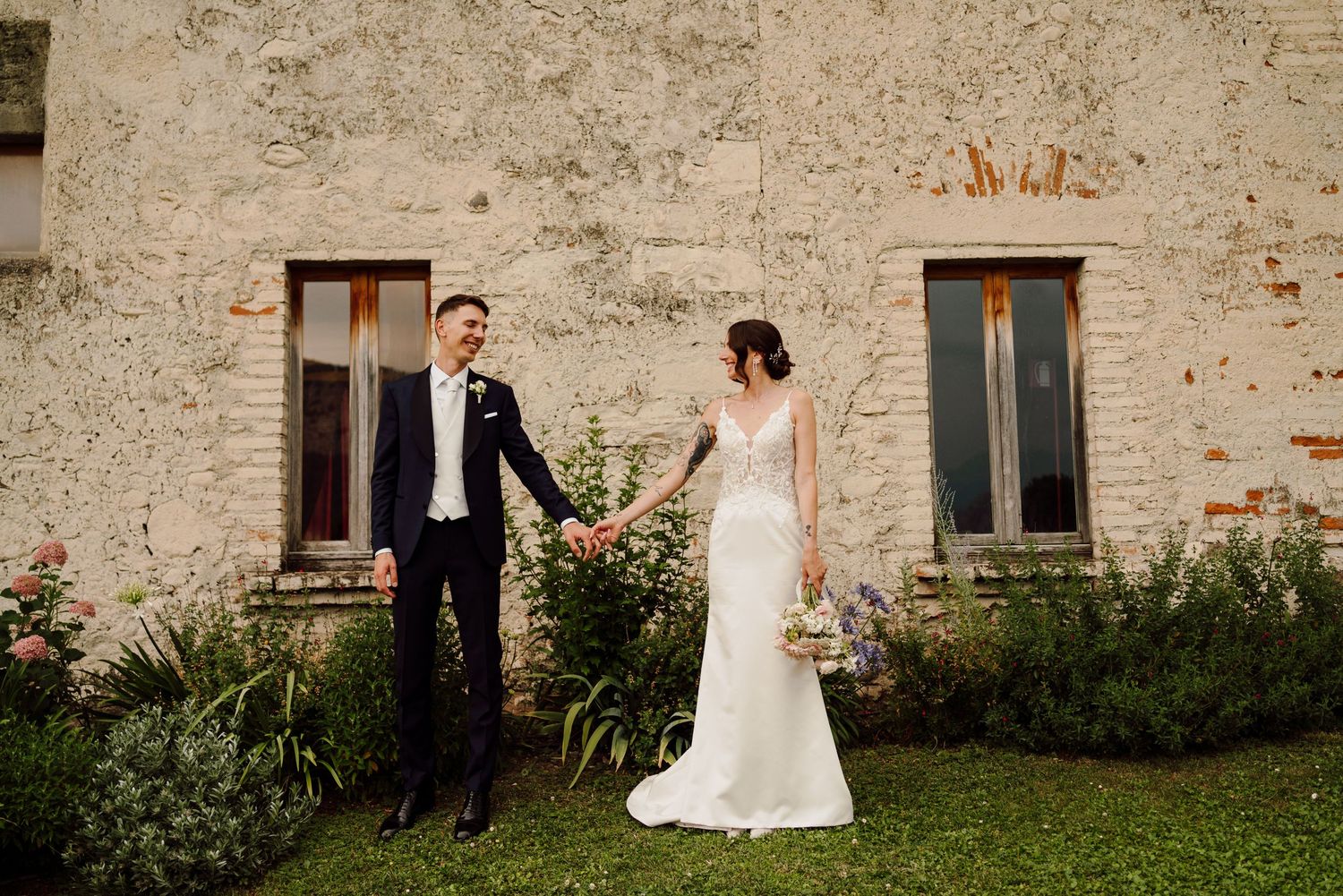 Bride and groom hold hands while walking beside a rustic stone building outdoors.