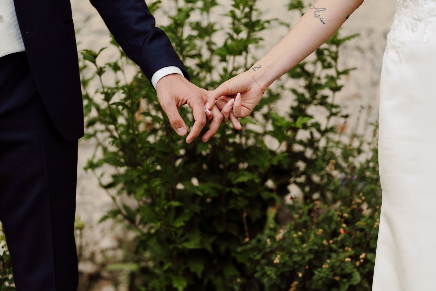 Close-up of bride and groom holding hands in front of blooming white flowers.