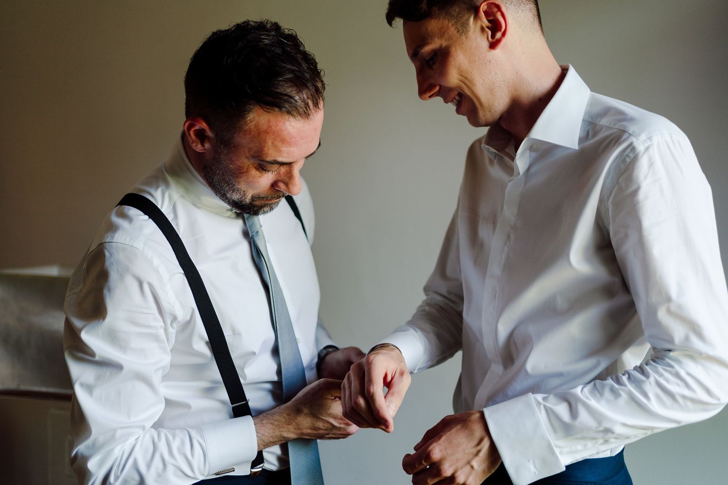 Two men in white shirts and suspenders helping each other with cufflinks during wedding preparation.