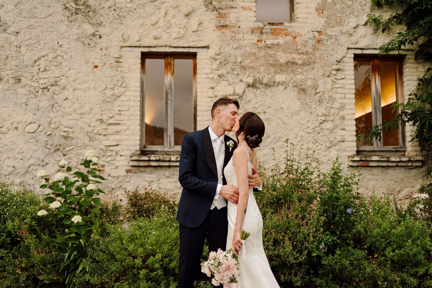 Bride and groom sharing a tender kiss beside a rustic stone building surrounded by lush greenery.