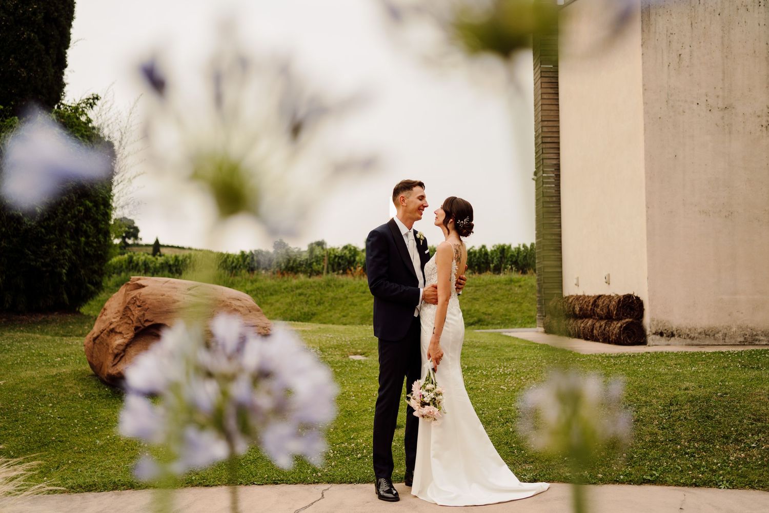 Bride and groom share a loving gaze framed by soft purple flowers in foreground.