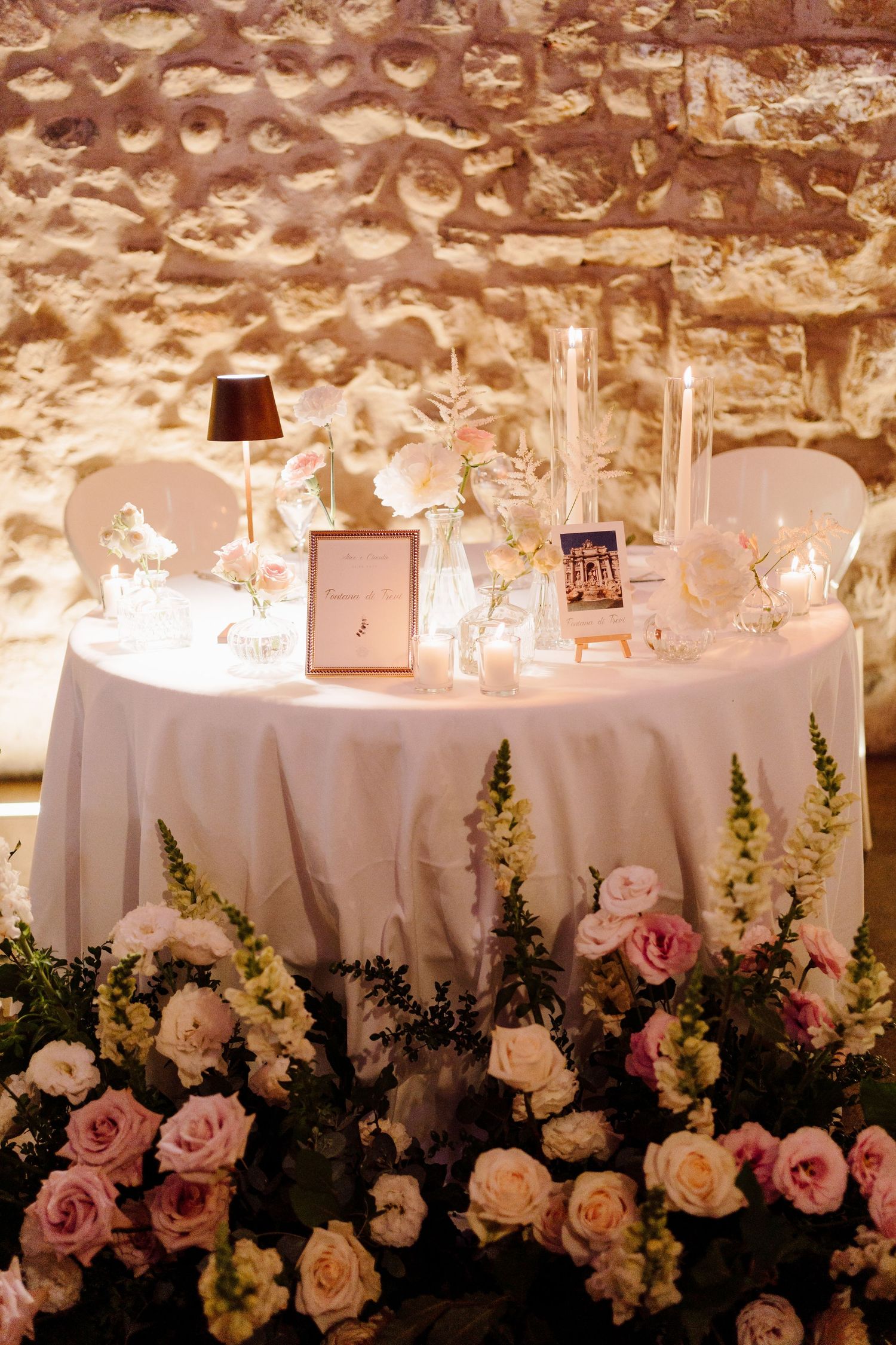 Romantic sweetheart table with white roses, pink flowers, candles, and gold frame against stone wall.
