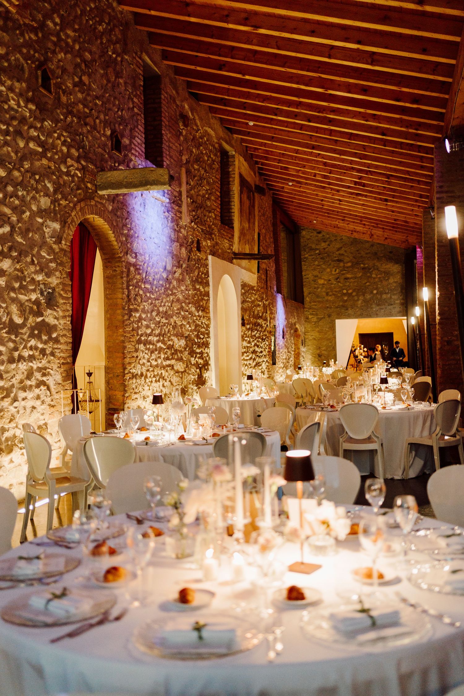 Grand wedding reception hall with stone walls, wooden ceiling, round tables, and warm candlelight.