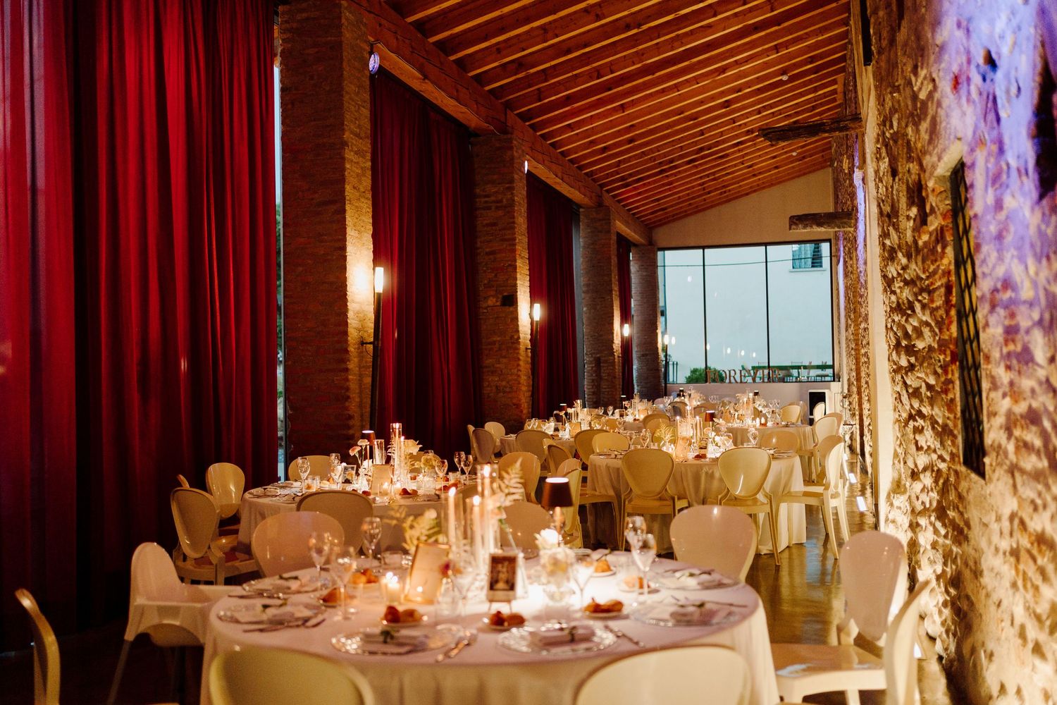 Intimate wedding reception room with red curtains, stone walls, candlelit round tables, and warm lighting.