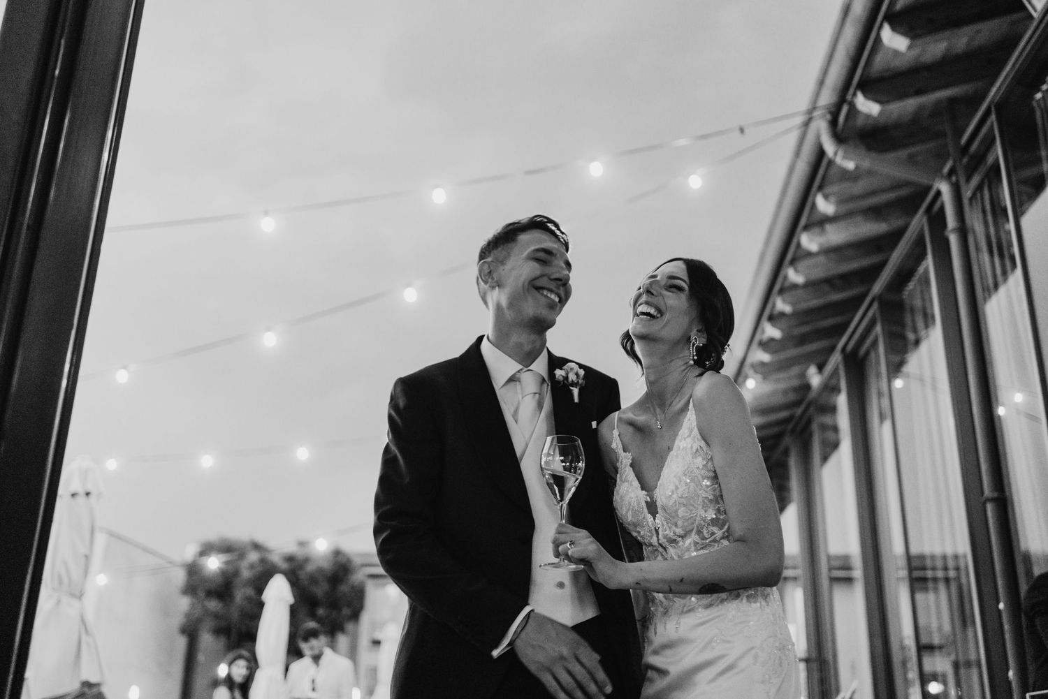 Happy bride and groom laughing under string lights in elegant black and white wedding portrait.