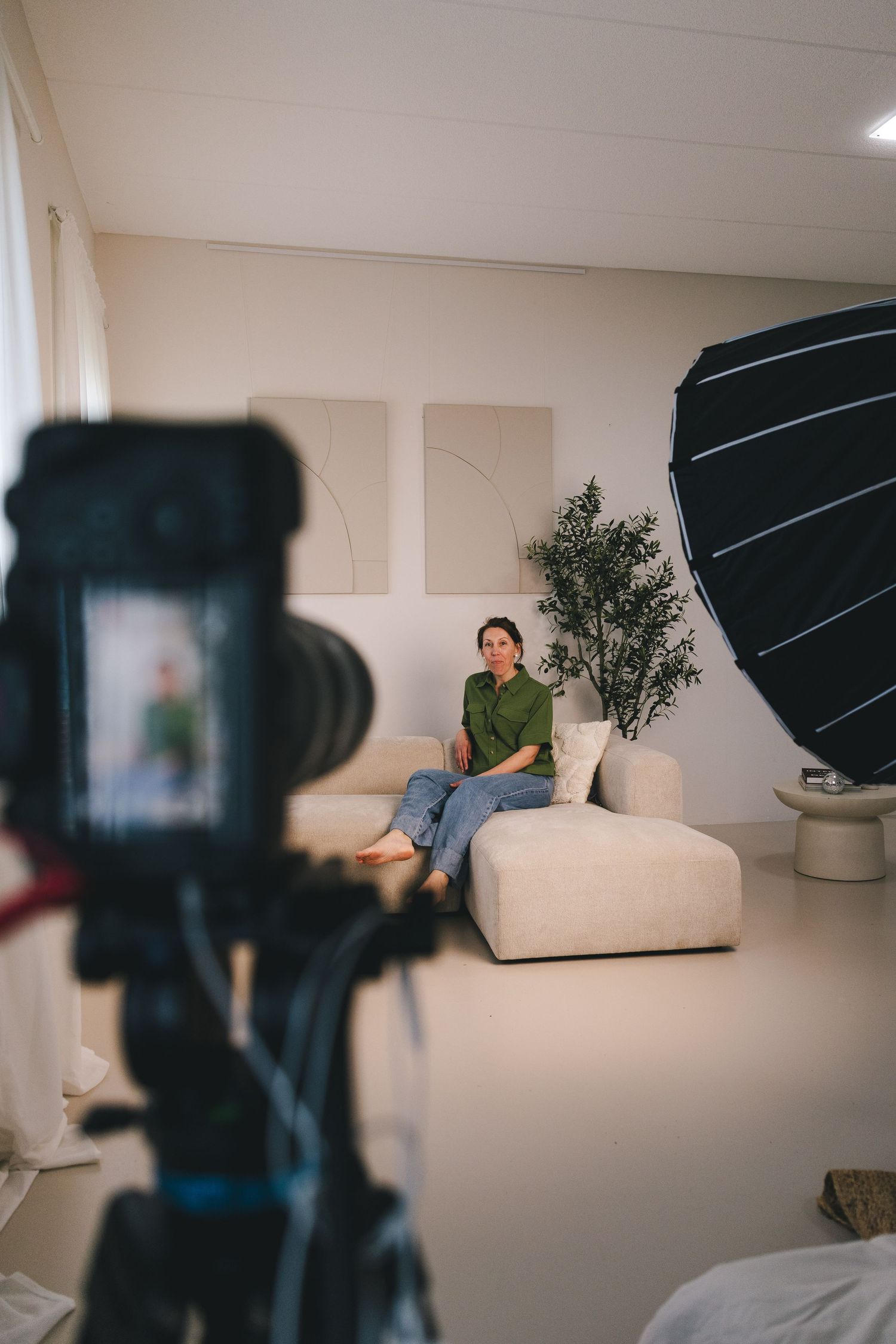 Woman in green shirt sits on white sofa in modern studio, photographed by camera on tripod with softbox light.