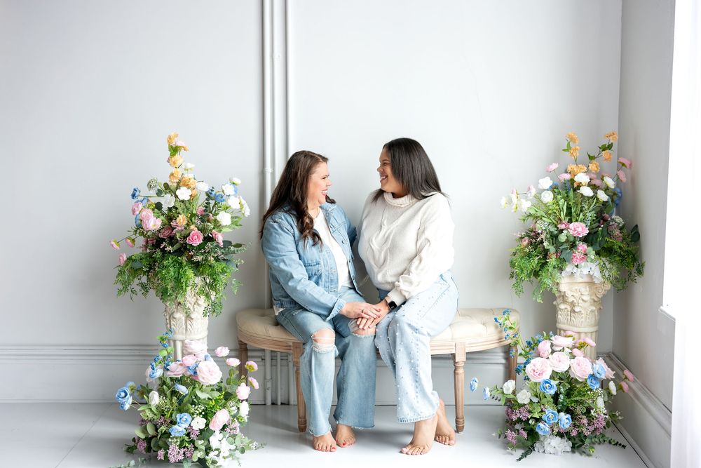 Two women in casual outfits hold hands on bench surrounded by pastel spring floral arrangements.