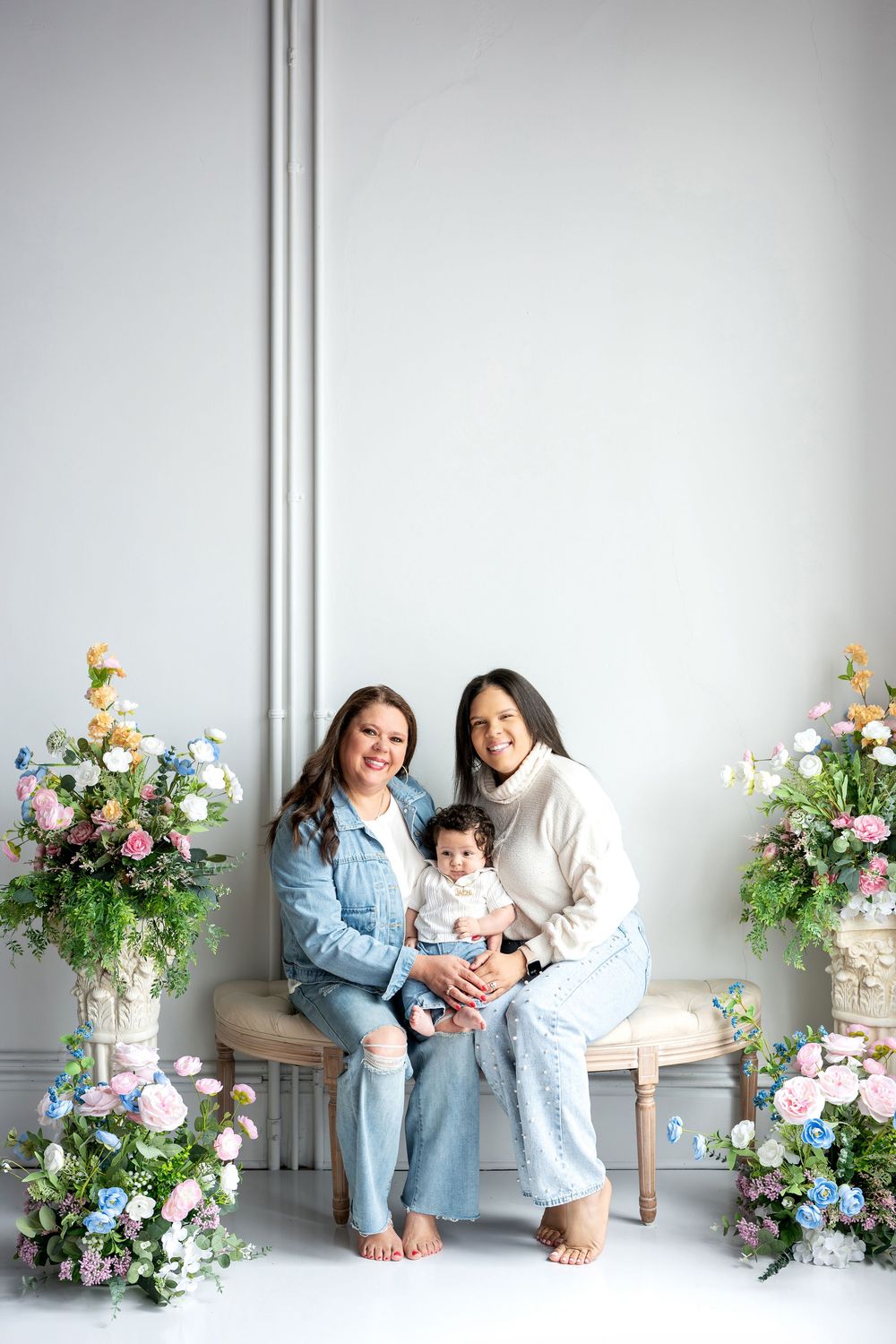 Two women and baby pose together on bench surrounded by colorful spring floral arrangements.