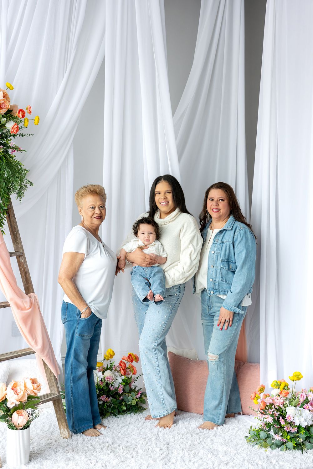 Three generations of women pose with a baby in a white draped studio with flowers.
