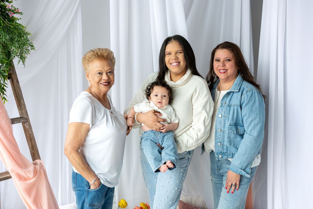 Three generations of women smile together holding a baby in a bright white studio.