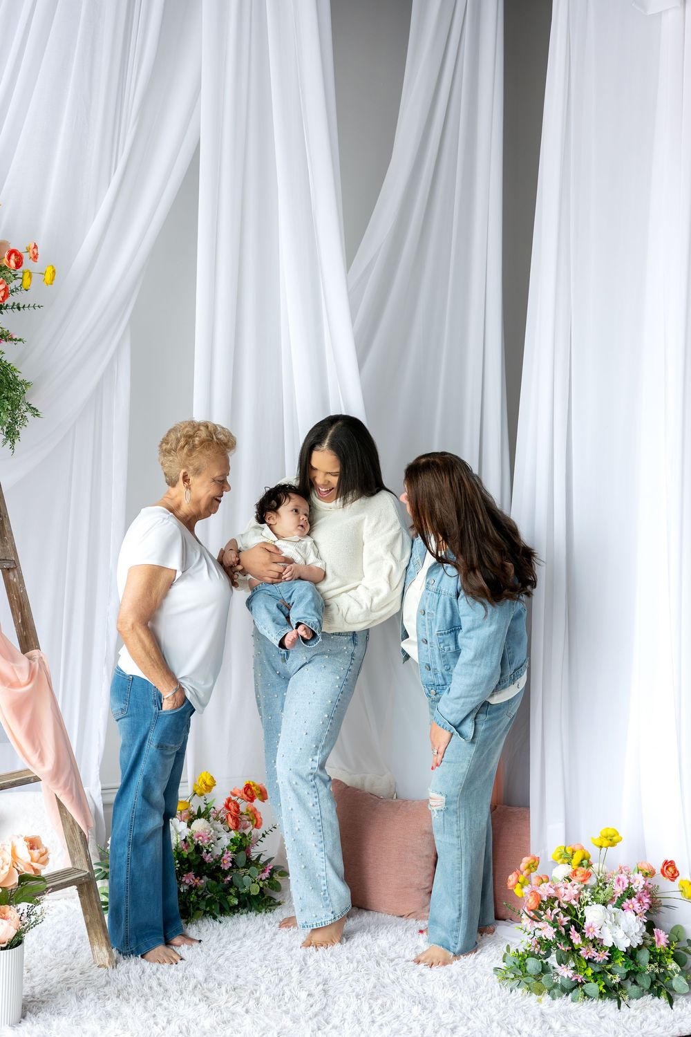 Three generations of women gather around a baby in a white studio with colorful flowers.