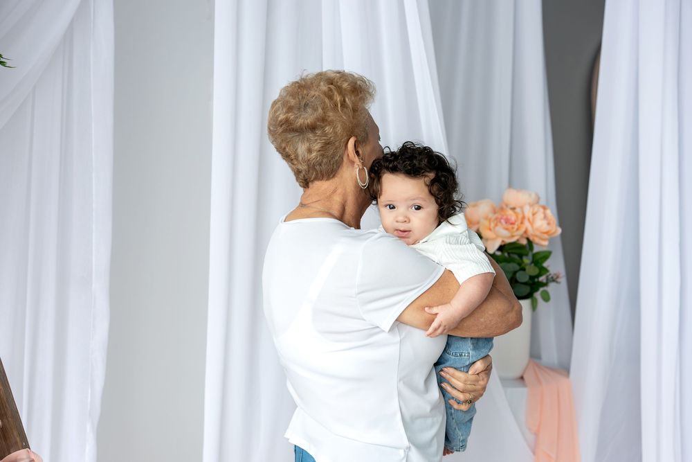 A grandmother holds a curly-haired baby near white drapes and peach roses.