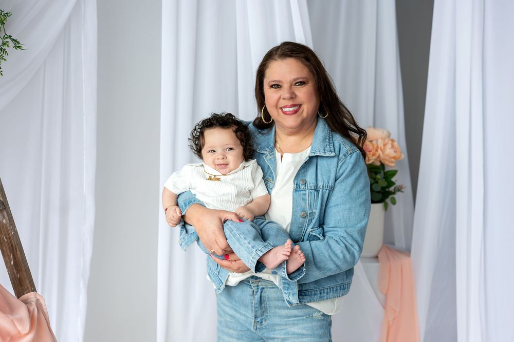 Smiling woman in denim jacket poses with curly-haired baby in white studio with floral decor.