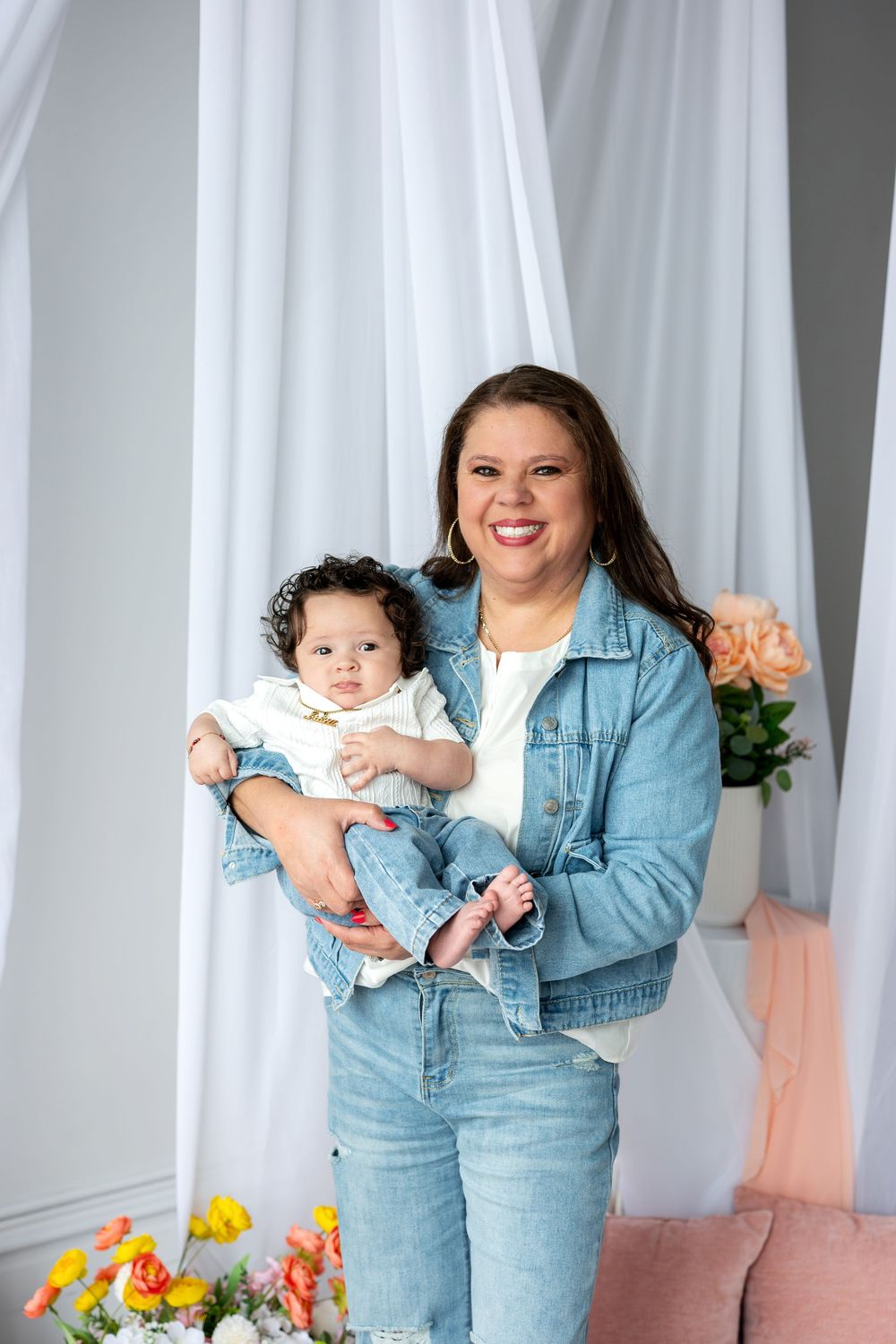 Happy woman in denim jacket holds baby girl in white onesie in bright floral photography studio.