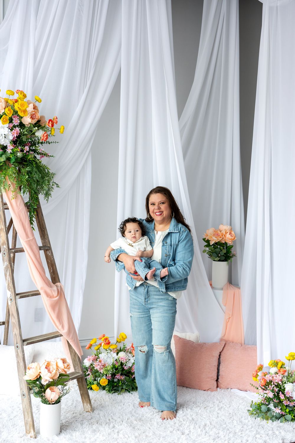 Woman in denim jacket holds baby while standing among colorful flowers and white draped backdrop.