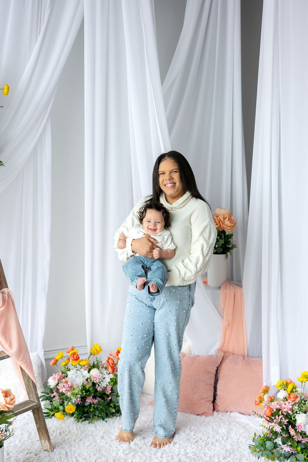 Smiling woman in cream sweater and blue pants holds curly-haired baby in bright floral studio.