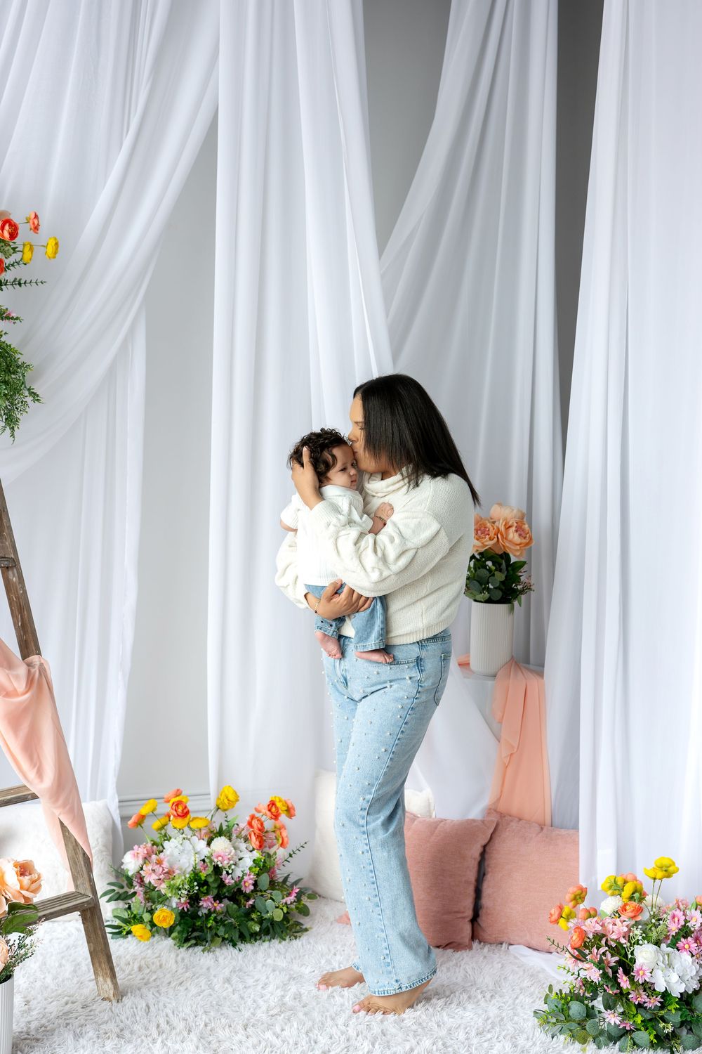Mother in cream sweater tenderly kisses baby while standing among colorful flowers and white drapes.