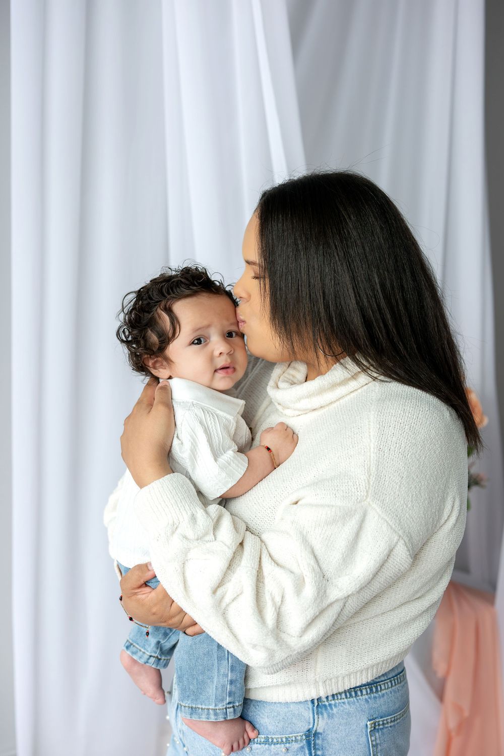 Young mother in white sweater tenderly kisses her curly-haired baby against white curtain backdrop.