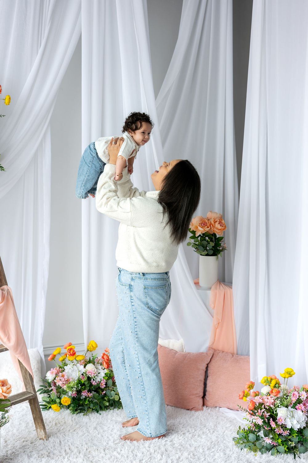 Joyful mother lifts smiling baby overhead in bright studio surrounded by colorful spring flowers.