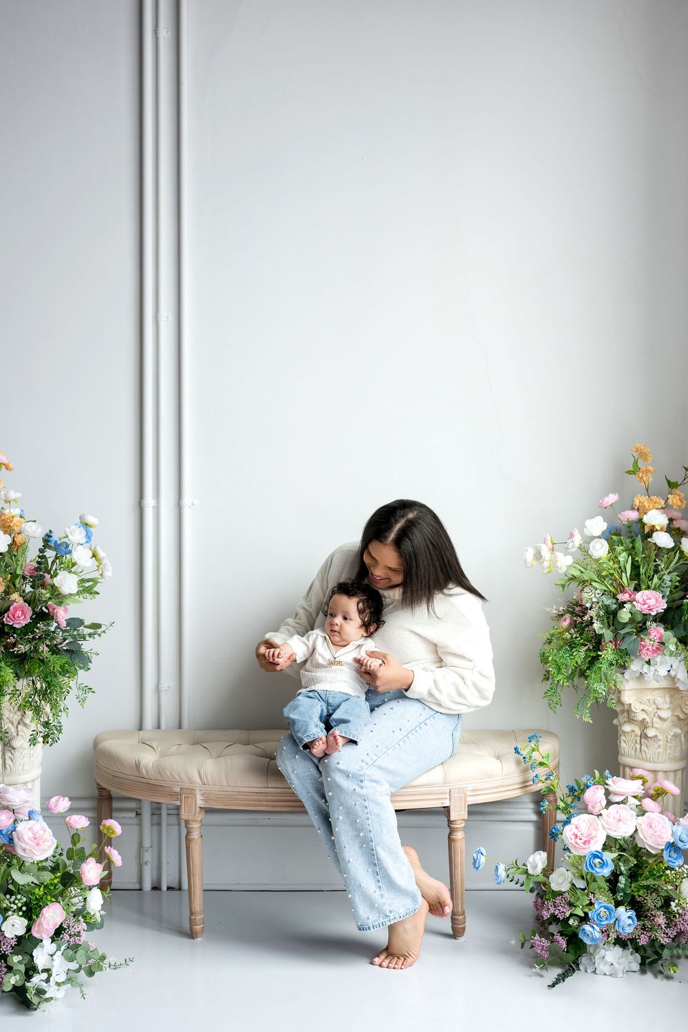 Mother in white sweater sits on vintage bench with baby surrounded by lush floral arrangements.