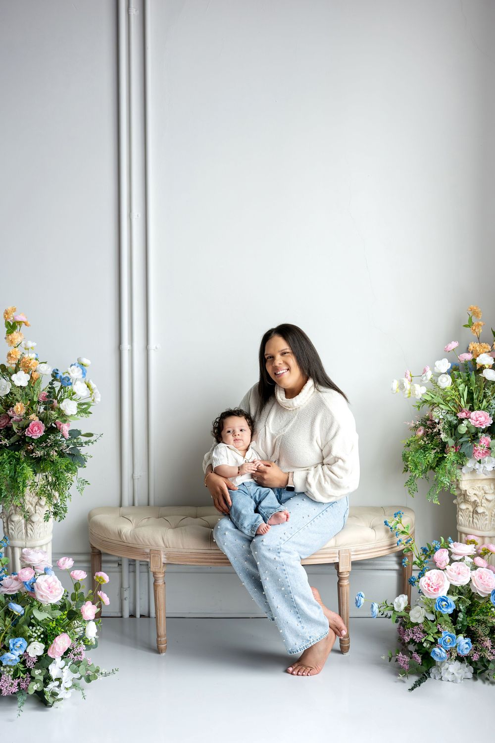 Smiling mother in white sweater holds baby on bench surrounded by colorful spring flowers.