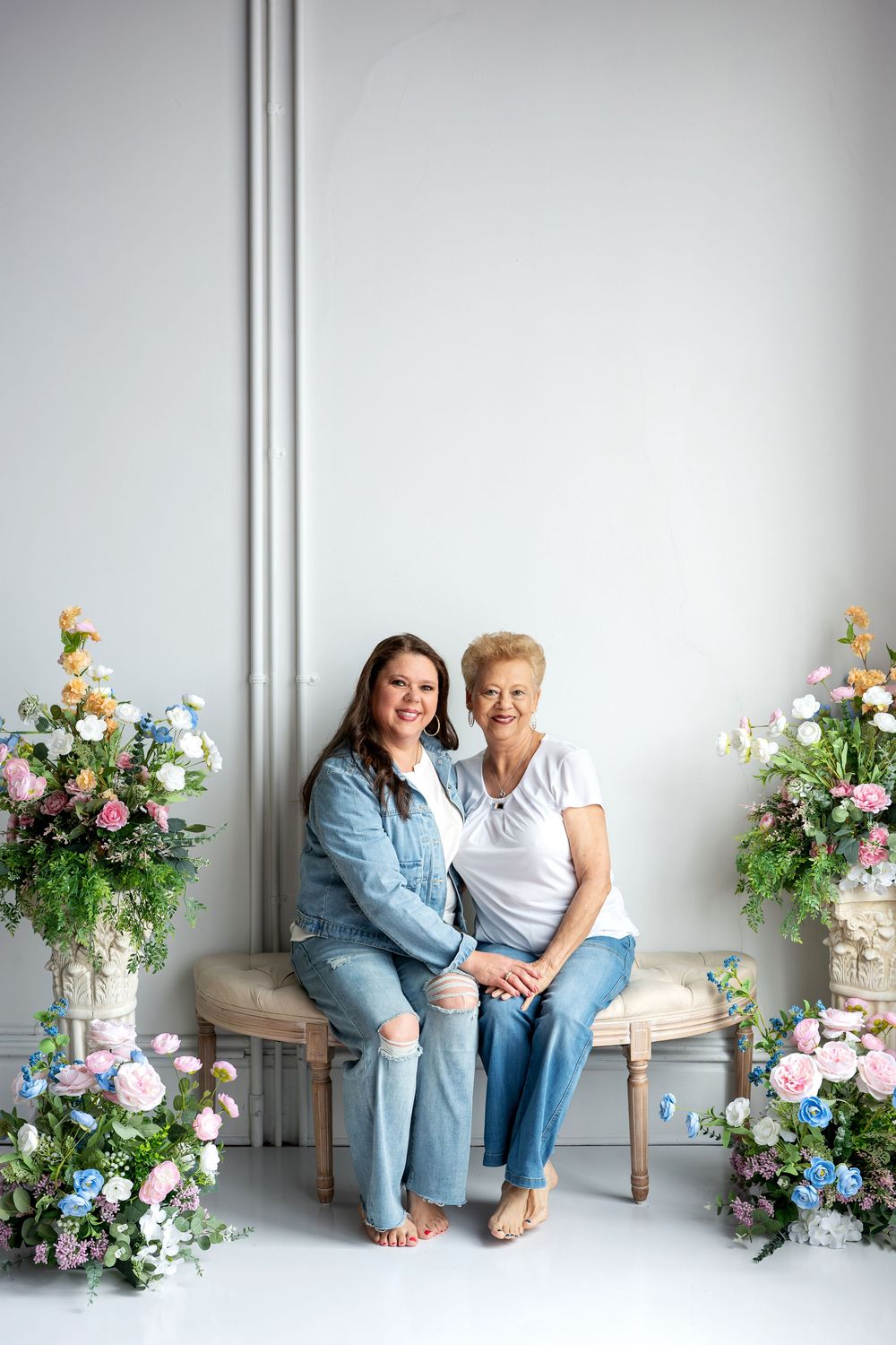 Two women in denim and white tops sit together on bench flanked by floral arrangements.